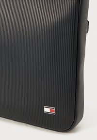 Black shoulder bag with ridged texture and smooth edges. Features a small logo with red, white, and blue accents on the front.