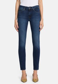 Schlanke blaue Jeans mit mittelhohem Bund, fünf Taschen und subtilen Verwaschungen, kombiniert mit gemusterten beigen Absätzen.