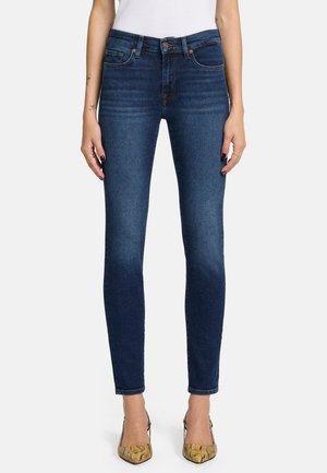 Jeans en denim bleu slim avec une taille mi-haute, un design à cinq poches et un léger délavage, associés à des talons beiges à motifs.