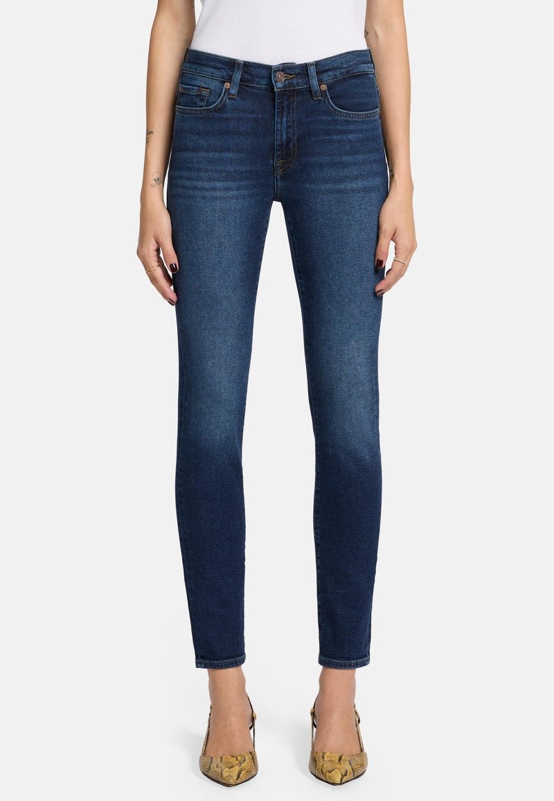 Schlanke blaue Jeans mit mittelhohem Bund, fünf Taschen und subtilen Verwaschungen, kombiniert mit gemusterten beigen Absätzen.