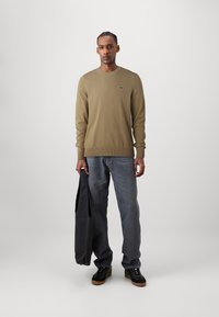 Olive green knitted sweater with round neckline, paired with dark gray jeans. The model holds a black bag. Black shoes with a thick sole.