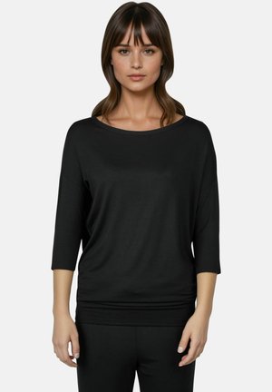 Vrouw met stijlbruin haar die een loszittend zwart driekwartsmouwen topje en zwarte broek draagt tegen een effen lichte achtergrond.