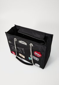Black fabric tote bag with silver hardware, two chain handles, interior zip pocket, and various colorful patches and branding details.