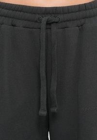 Pantalons de survêtement noirs avec une taille à cordon de serrage, présentant une texture lisse et des poignets élastiques. Le logo de la marque est subtilement imprimé sur le côté.