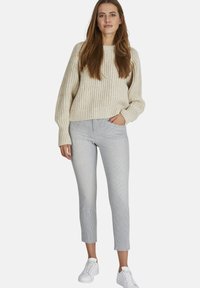 Beige gebreide trui met een losse pasvorm, gecombineerd met slim-fit broek in grijs en wit met strepen en witte sneakers. Casual outfit met textuurdetails.