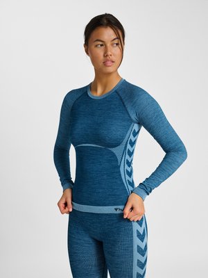 Junge Frau, die ein blaues Langarm-Sportoberteil mit gemusterten Seitenteilen anpasst, vor einem schlichten hellen Hintergrund stehend.