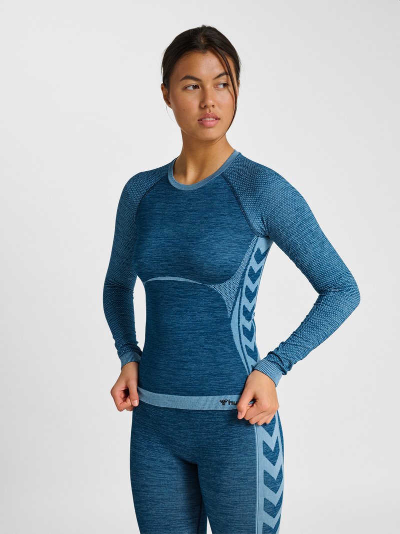 Junge Frau, die ein blaues Langarm-Sportoberteil mit gemusterten Seitenteilen anpasst, vor einem schlichten hellen Hintergrund stehend.