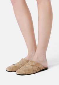 Mules de ante en beige con un diseño tejido de patrones en diamante. Suela negra de perfil bajo y parte trasera abierta para un fácil uso.