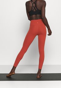 Punatut leggingsit sileästä, joustavasta materiaalista korkean vyötärön kanssa, yhdistettynä mustaan nauhamalliseen urheiluliiviin, nähtynä takaa.