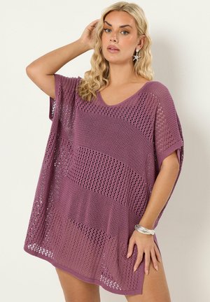 Lyshåret kvinde iført lilla løst strikket overtræk, søstjerneøreringe, sølvarmbånd, poserer med hånden nær hovedet mod en hvid væg.