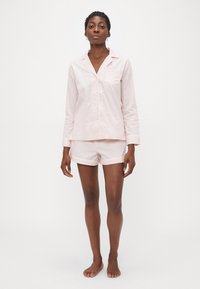 Kvinne som står barfot iført en lys rosa langermet skjorte med knapper og matchende shorts pysjamas mot en enkel hvit bakgrunn.