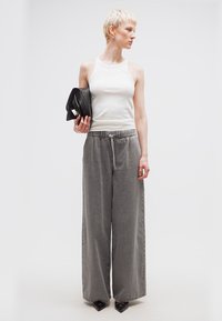 Άτομο με κοντά ξανθά μαλλιά που φοράει λευκό αμάνικο τοπ, γκρι παντελόνι με φαρδιά μπατζάκια και μαύρα μυτερά παπούτσια, κρατώντας μια μαύρη τσάντα χειρός, κοιτάζοντας δεξιά.