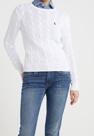 Person iført en hvid kabelstrikket sweater over en blå skjorte med krave og blå jeans, stående med den ene hånd i lommen.