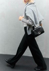Grauer gestrickter Überwurf über einem gestreiften Langarmshirt, kombiniert mit weiten schwarzen Hosen und schwarzen Ballerinas. Als Accessoires dient eine kleine schwarze Handtasche.