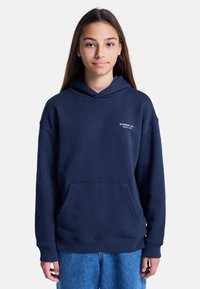 Chica adolescente con cabello largo castaño, vistiendo una sudadera azul marino con el texto "ELEMENT CO. SINCE 1992" y jeans azules, de pie contra un fondo blanco.