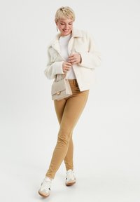 Veste en fausse fourrure crème, haut à manches longues blanc, pantalon beige ajusté et baskets blanches. Sac à main beige simple avec un détail de fermoir doré.