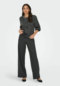 Tumedast hall kaelusega kudum, millel on nupud ja kaunistused, koos mustade ja valgete triibuliste avarate pükstega ning tekstureeritud mustade lamedate kingadega.