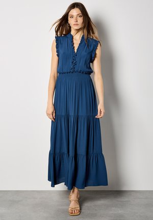 Femme debout contre un mur blanc, portant une robe maxi bleu sans manches avec des volants et des sandales beiges à lanières.