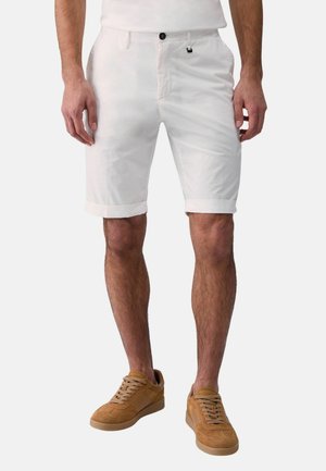 Uomo che indossa pantaloncini bianchi al ginocchio e sneakers in camoscio color beige su uno sfondo semplice.