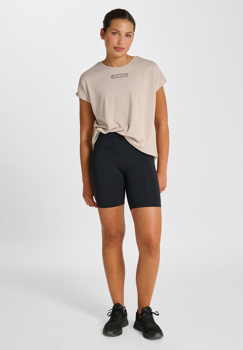 Beige sportt-shirt med logotyp, lös passform, korta ärmar, kombinerad med svarta cykelbyxor med hög midja och svarta sportskor.