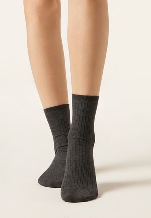 Gambe con calzini grigi scuri a coste fino a metà polpaccio, in piedi su una superficie chiara con uno sfondo chiaro e uniforme.