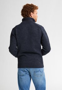 Bărbat cu păr cârlionțat, îmbrăcat cu un pulover negru închis cu gât și blugi de culoare bleu deschis, stând cu fața întors către un fundal simplu.