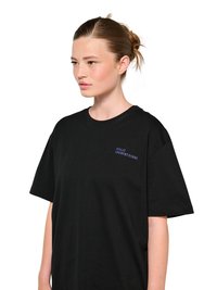 Jeune femme à la peau claire et aux cheveux bruns attachés, portant un t-shirt noir ample avec un petit texte bleu sur la poitrine, tournée vers la gauche.