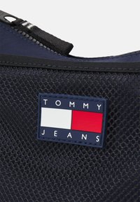 Navy mesh fabric with a black zipper, featuring a multicolored logo patch with blue, white, and red rectangles, and 'TOMMY JEANS' text.