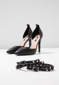 Chaussures à talons hauts noires à bouts pointus placées derrière deux sangles en cuir noir ornées de boucles en argent et de clous métalliques circulaires.