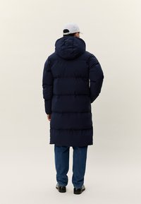 Persona, kas vērsta prom, valkājot tumši zilu garu pūkainu mēteli ar kapuci, zilas džinsas, melnas kurpes un gaiši pelēku beisbola cepuri.