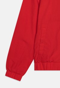 Veste rouge à manches longues avec poignets et bas élastiques, dotée d’une poche fendue verticale sur le côté, sur un fond clair uni.