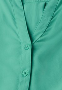 Blouse boutonnée en tissu uni teal. Présente une texture douce, un col en V et des fermetures à boutons avec un accent de couleur assortie.