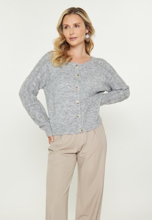 Vaaleanharmaa neulecardigan, jossa on pyöreä kaula-aukko, koristeellisia nappeja rivissä ja ribbauskohdat hihoissa, kerroksittain beigen värisien housujen päällä.