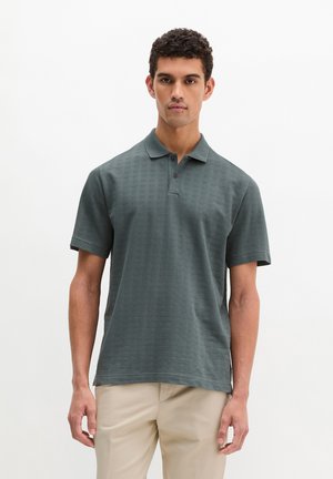 Młody mężczyzna w ciemnozielonej koszulce polo z krótkim rękawem i beżowych spodniach, stojący na jednolitym białym tle.