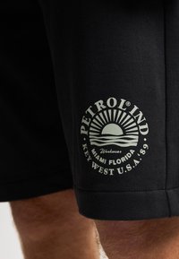 Pantalones cortos negros de algodón con un diseño ajustado y elegante. Presentan un gráfico impreso de un sol en color claro con el texto: "PETROL IND, MIAMI FLORIDA."