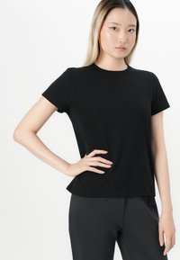 Jeune femme avec des cheveux blonds lisses portant un t-shirt noir uni et un pantalon sombre, posant avec une main sur la hanche devant un fond blanc.