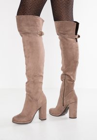 Botas de ante hasta el muslo en tono topo, con un tacón bloque, cremallera lateral y un hebilla decorativa en la parte superior. Textura suave y diseño ajustado.