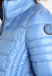 Himmelblau, gesteppte Daunenjacke mit hohem Kragen, ausgestattet mit einem runden Logopatch am Oberarm und einer glatten, glänzenden Textur.