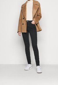 Abrigo de mezcla de lana beige de doble botonadura con grandes solapas, combinado con jeans skinny negros y zapatillas blancas. Texturas suaves, corte ajustado.