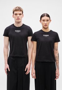 T-shirts pretas de manga curta em algodão com o logótipo "TOMMY JEANS" em branco, usadas por duas pessoas, combinadas com calças largas pretas.