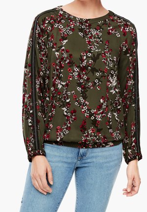 Kobieta ubrana w oliwkową bluzkę z długim rękawem w czerwono-biały kwiatowy wzór, zestawioną z jasnoniebieskimi dżinsami, pokazana od pasa w górę wraz z rękami.