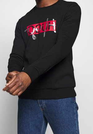 Persona que lleva un sudadera negra de manga larga con el logo "HUGO" en rojo y plata invertido, combinado con unos pantalones de mezclilla azules.