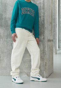 Sweatshirt teal avec "DICKIES" imprimé sur la poitrine ; pantalon blanc texturé ; baskets blanches avec des accents verts ; posé contre un mur en béton.