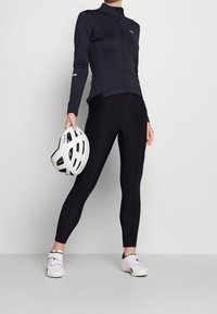 Black fitted leggings paired with a long-sleeve zip-up top, both made of stretch fabric. A white cycling helmet is held in one hand.