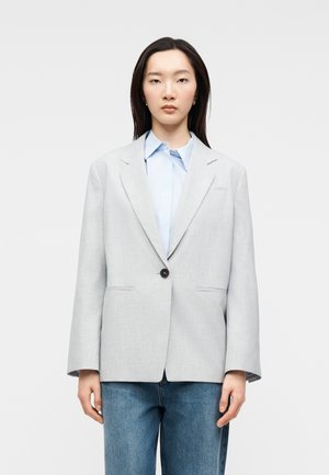 Jonge vrouw staande, gekleed in een oversized lichtgrijze blazer over een lichtblauw overhemd met kraag en blauwe spijkerbroek tegen een effen achtergrond.