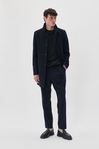 Marineblå uldfrakke med knaplukninger, parret med en sort sweater og marineblå bukser. Modellen har sorte loafers og lysegrå sokker på.