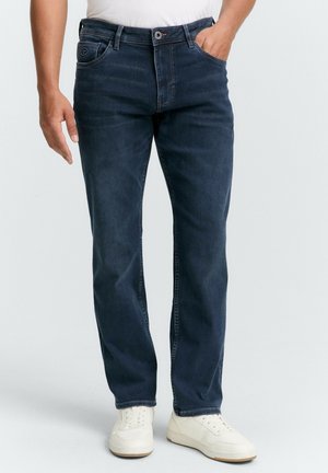 Homme portant un jean droit bleu foncé et des baskets blanches, debout avec une main dans la poche devant un fond uni.