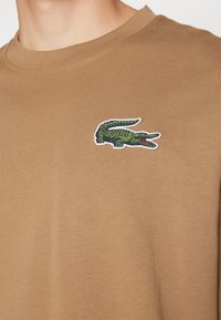 T-shirt in cotone marrone con logo di un coccodrillo verde ricamato sul petto, col collo tondo classico e texture liscia.
