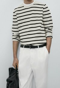 Gestreifter Strickpullover in Creme und Schwarz, Rundhalsausschnitt, lange Ärmel. Kombiniert mit weißen Hosen und einem schwarzen Gürtel. Schwarze Handtasche in der Hand.