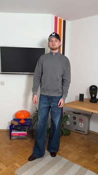 Gray zip-up jacket, blue wide-leg jeans, and black shoes. Accessories include a black cap and tattoos on the forearm. Modern interior setting.
